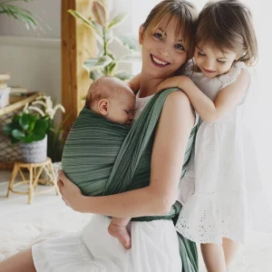 Madre sentada en una alfombra blanca sonriendo con su bebé dormido en el fular tejido verde Verdelite mientras la hermana mayor la abraza por el cuello.