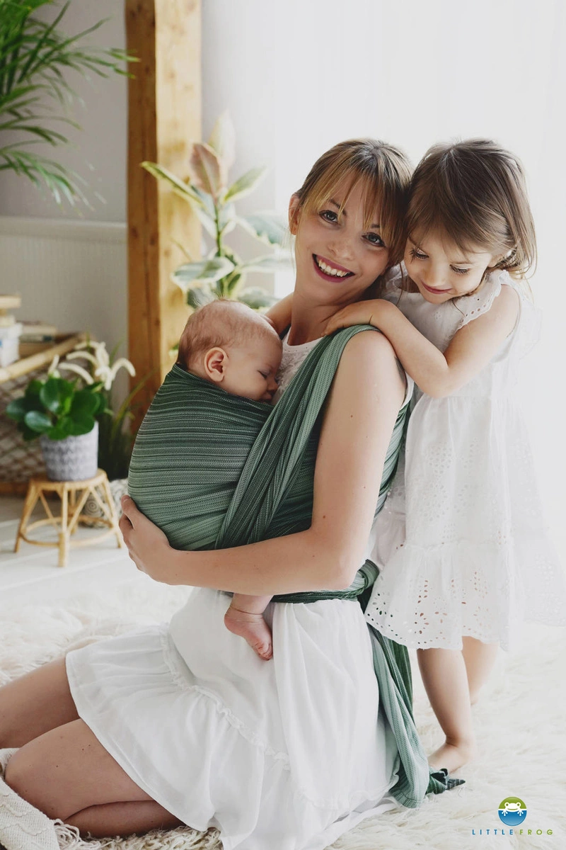 Madre sentada en una alfombra blanca sonriendo con su bebé dormido en el fular tejido verde Verdelite mientras la hermana mayor la abraza por el cuello.