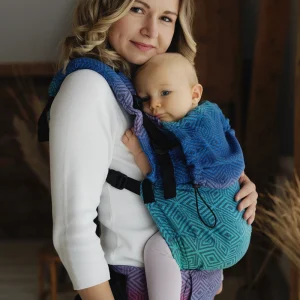Madre sonriendo de perfil portando a su bebé en la mochila ergonómica evolutiva Little Frog Prime V3 con tejido de fular geométrico en tonos azules, turquesas y morados Aurora Cube.