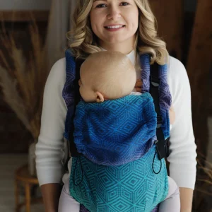 Madre sonriendo de frente sujetando la base de la mochila evolutiva Little Frog Prime V3 Aurora Cube mientras portea a su bebé en postura ergonómica piernas en M