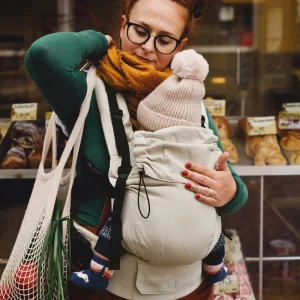 Madre ajustando el tirante de la mochila evolutiva beige Little Frog Prime V3 mientras sostiene a su bebé y lleva una bolsa de la compra.