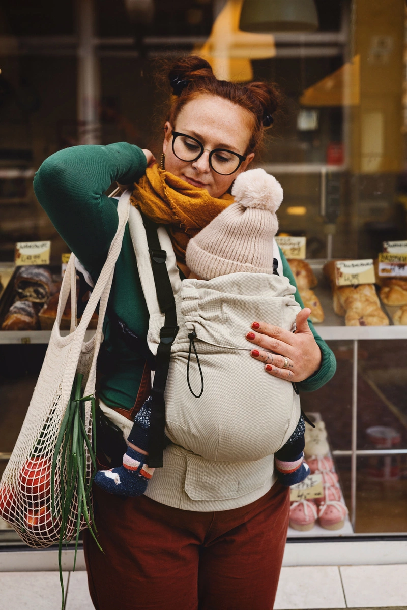 Madre ajustando el tirante de la mochila evolutiva beige Little Frog Prime V3 mientras sostiene a su bebé y lleva una bolsa de la compra.