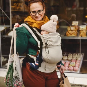 Madre de pie sonriendo a cámara con su bebé porteado en la mochila evolutiva beige Little Frog Prime V3 sosteniendo bolsas de la compra.