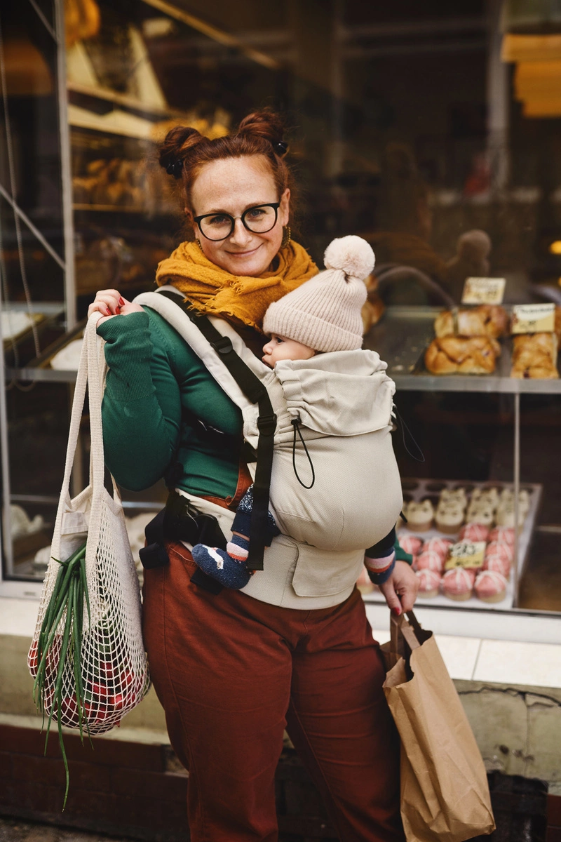 Madre de pie sonriendo a cámara con su bebé porteado en la mochila evolutiva beige Little Frog Prime V3 sosteniendo bolsas de la compra.
