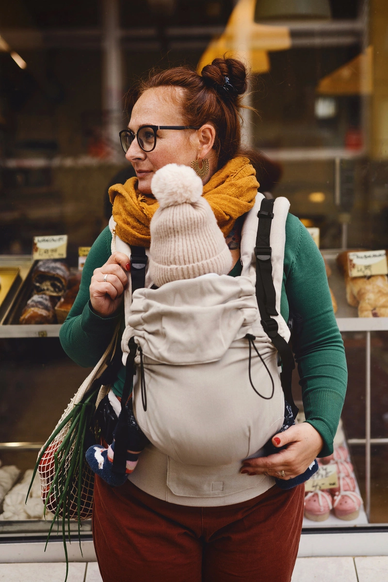 Plano medio de madre mirando al frente con su bebé porteado ergonómicamente en la mochila evolutiva Little Frog Prime V3 color beige.