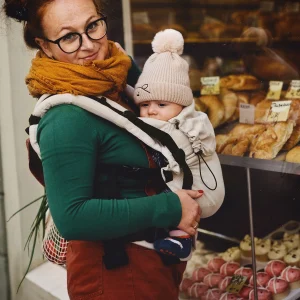 Madre sonriendo de tres cuartos frente a la panadería con su bebé bien abrigado en la mochila evolutiva Little Frog Prime V3 Beige