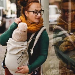 Madre observando un escaparate de perfil con su bebé porteado en la mochila evolutiva beige Little Frog Prime V3.