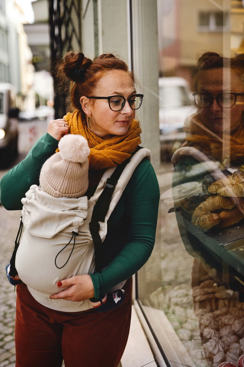 Madre observando un escaparate de perfil con su bebé porteado en la mochila evolutiva beige Little Frog Prime V3.