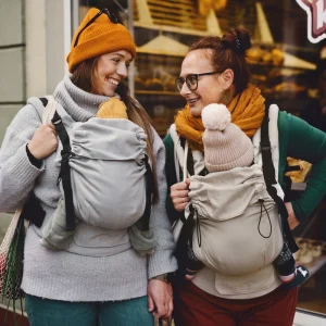 Dos madres paseando juntas por la calle porteando a sus bebés en mochilas evolutivas Little Frog Prime V3 en tonos gris y beige.