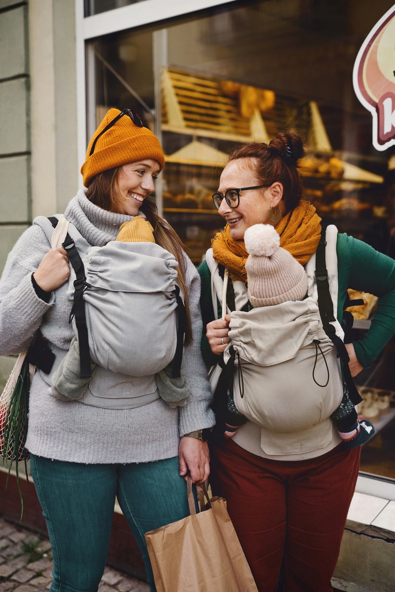 Dos madres paseando juntas por la calle porteando a sus bebés en mochilas evolutivas Little Frog Prime V3 en tonos gris y beige.