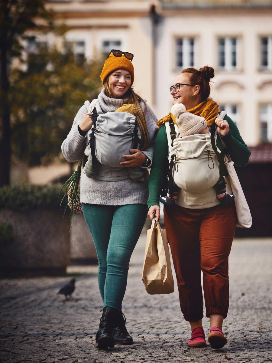 Dos madres caminando por una calle adoquinada conversando y porteando a sus bebés con mochilas evolutivas Little Frog Prime V3.