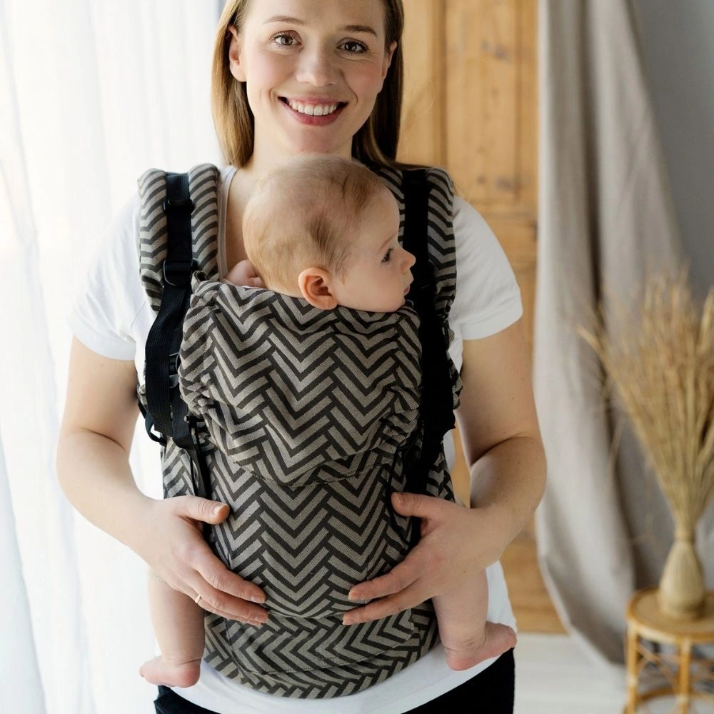 Plano medio de madre sonriendo con su bebé mirando de perfil en la mochila evolutiva de fular Little Frog Prime Dragon Miles.