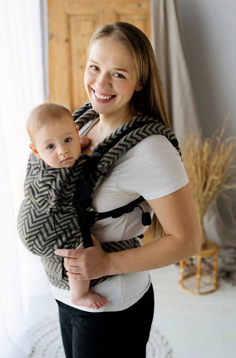 Madre sonriendo de perfil junto a la ventana mientras portea a su bebé en la mochila evolutiva Little Frog Prime Dragon Miles en tonos negro y beige con patrón en zigzag.