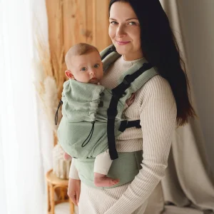 Madre sonriendo de pie mirando a cámara portando a su bebé al frente con la mochila ergonómica evolutiva verde salvia Little Frog Prime V3.