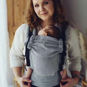 Madre mirando a cámara sonriendo dulcemente mientras portea a su recién nacido en la mochila evolutiva Little Frog Prime Zen Path con patrón en espiga gris y blanco.