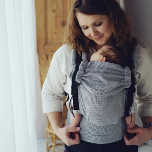 Madre mirando con ternura a su bebé recién nacido dormido en la mochila ergonómica evolutiva Little Frog Prime Zen Path en tono gris espiga.