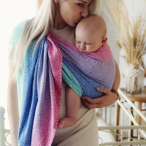 Madre rubia besando la cabeza de su bebé dormido, porteado ergonómicamente en una bandolera de anillas multicolor Little Frog Aurora Cube de algodón peinado.