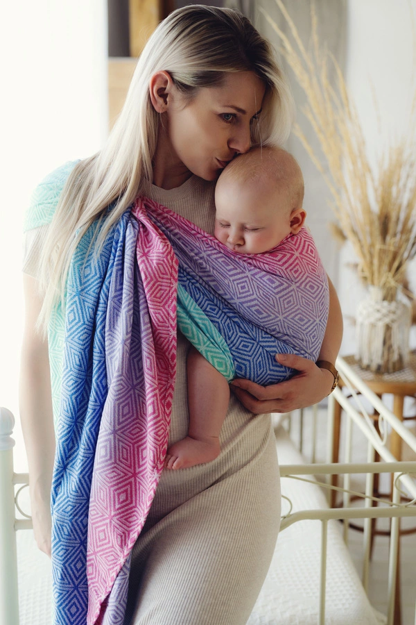 Madre rubia besando la cabeza de su bebé dormido, porteado ergonómicamente en una bandolera de anillas multicolor Little Frog Aurora Cube de algodón peinado.