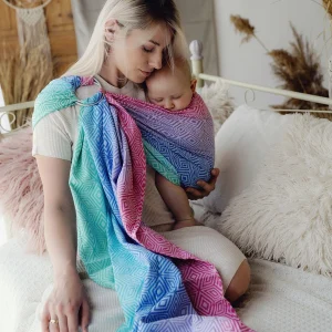 Madre sentada en una cama blanca mirando con ternura a su bebé dormido en la bandolera de anillas multicolor Little Frog Aurora Cube.
