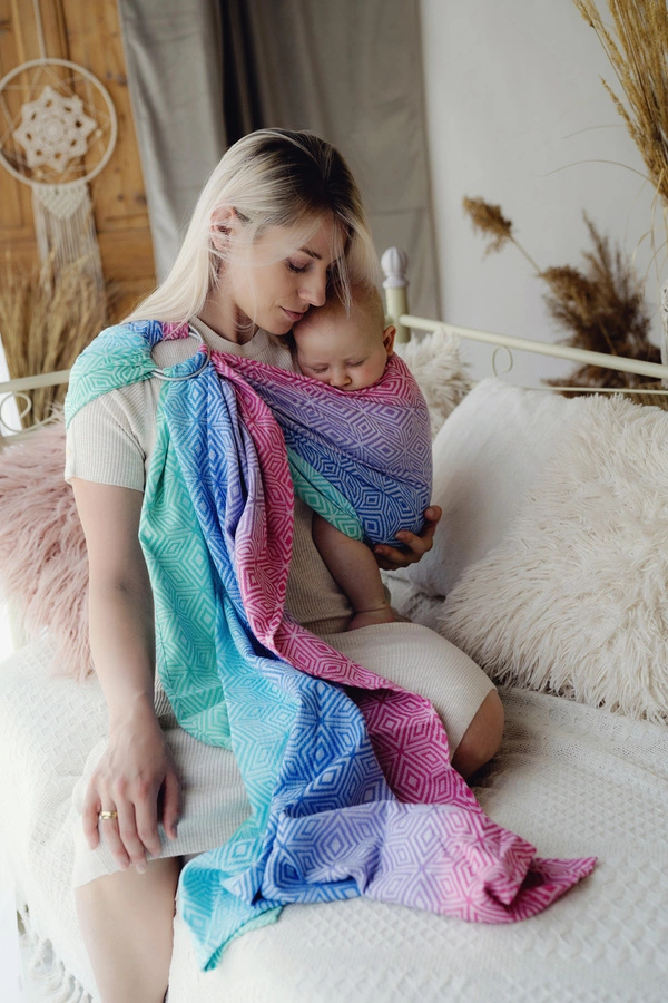 Madre sentada en una cama blanca mirando con ternura a su bebé dormido en la bandolera de anillas multicolor Little Frog Aurora Cube.