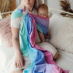 Madre sentada en una cama de tonos neutros porteando a su bebé en la bandolera de anillas Little Frog Aurora Cube con diseño de cubos en tonos rosa, azul y turquesa.