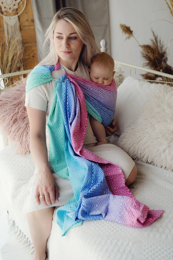 Madre sentada en una cama de tonos neutros porteando a su bebé en la bandolera de anillas Little Frog Aurora Cube con diseño de cubos en tonos rosa, azul y turquesa.