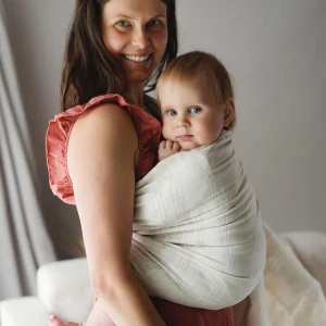 Madre sonriente con vestido color teja sosteniendo la piernita de su bebé de ojos azules alojado ergonómicamente en la bandolera de anillas de lino blanco Little Frog Summer Meadow.