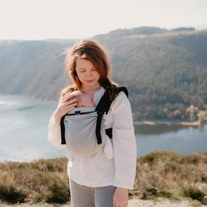 Madre caminando al aire libre observando a su recién nacido dormido ergonómicamente alojado en la mochila evolutiva Néobulle My Néo Grey con tirantes negros.