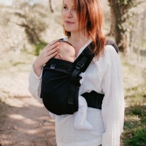 Madre con camisa blanca acariciando a su recién nacido dormido ergonómicamente alojado en la mochila evolutiva Néobulle My Néo totalmente negra.