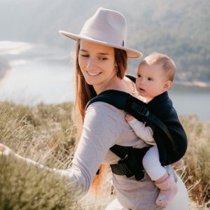 Madre con sombrero beis sonriendo a su bebé porteado a la espalda en la mochila ergonómica Néobulle My Néo Noir frente a un paisaje natural.