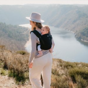 Plano medio de madre con sombrero porteando a su bebé a la espalda con la mochila ergonómica Néobulle My Néo Black en un mirador con vistas a un río.