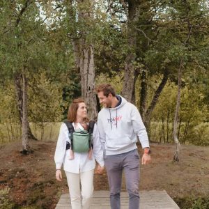 Pareja caminando de la mano por un sendero arbolado mientras la madre portea a su recién nacido en la mochila evolutiva Néobulle My Néo Vert.